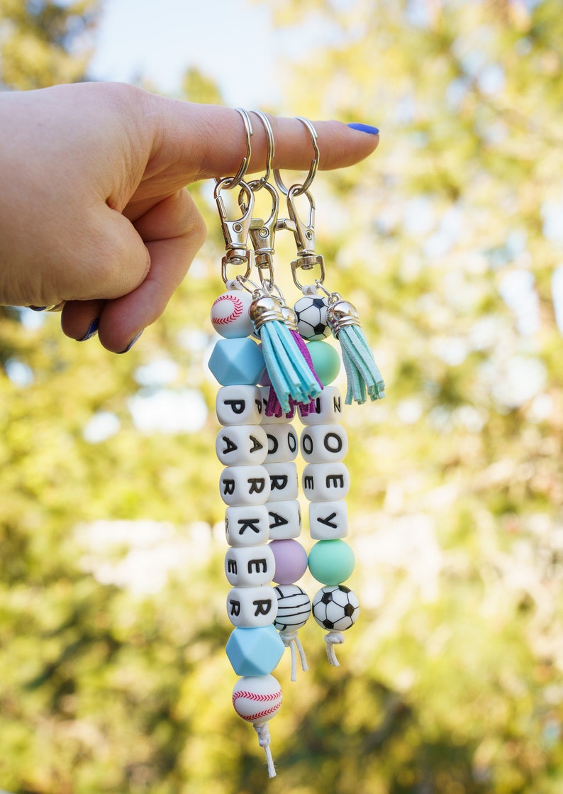 Pu&ograve; includere: Tre portachiavi personalizzati con chiusure argentate, ognuno con un nome scritto con perline di lettere bianche. I portachiavi includono perline decorative di vari colori e ciondoli a tema sportivo.
