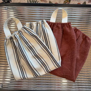 May include: Three reusable fabric produce bags. One bag is cream-colored with black vertical stripes, and the other two are a solid reddish-brown color. Each bag has a white fabric handle.