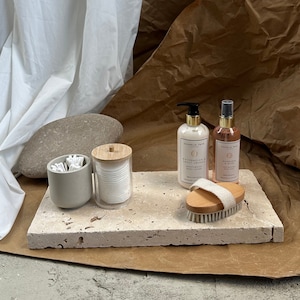 May include: A light brown rectangular tray with a white, textured surface. On the tray are two bottles of body lotion, a small container of cotton rounds, a container of cotton swabs, and a wooden brush with a white strap.