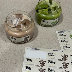 Op de afbeelding: Twee glazen potkaarsen, &eacute;&eacute;n met lichtbruine was en de tekst "ICED LATTE", de andere met groene was en de tekst "ICED MATCHA". Beide kaarsen hebben ijsblokjes bovenop. Stickers met de tekst "ICED LATTE" en "ICED MATCHA" zijn ook zichtbaar.