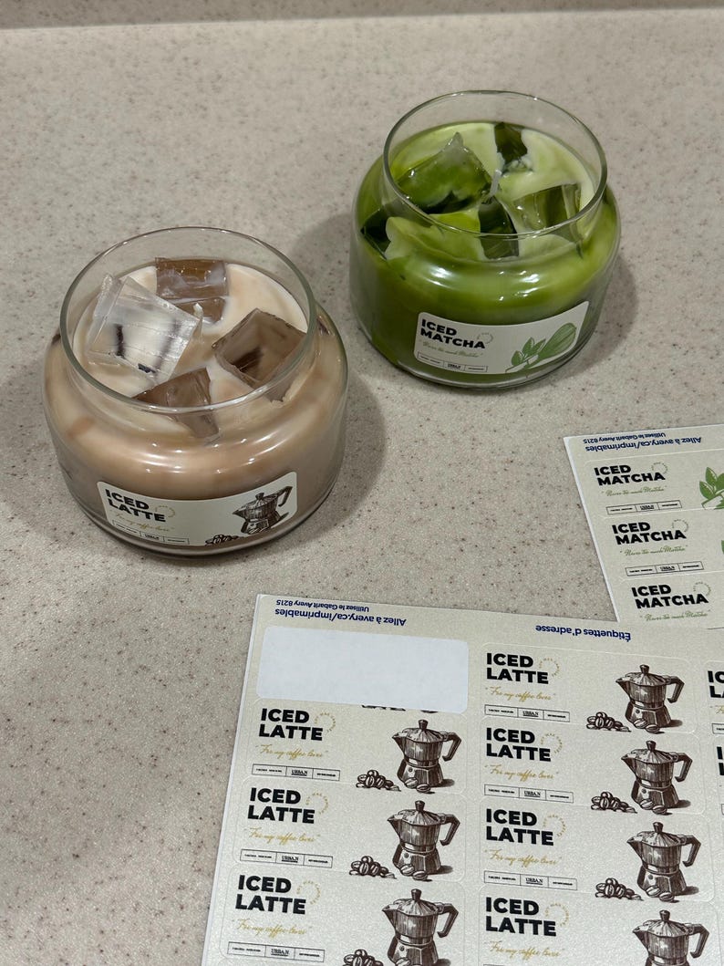 Op de afbeelding: Twee glazen potkaarsen, &eacute;&eacute;n met lichtbruine was en de tekst "ICED LATTE", de andere met groene was en de tekst "ICED MATCHA". Beide kaarsen hebben ijsblokjes bovenop. Stickers met de tekst "ICED LATTE" en "ICED MATCHA" zijn ook zichtbaar.