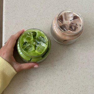 Op de afbeelding: Twee ijskoude drankjes in doorzichtige glazen potten. E&eacute;n is een felgroene matcha latte met ijsblokjes, de andere een lichtbruine koffiedrank met ijs. De drankjes staan op een gespikkeld, lichtgekleurd oppervlak.
