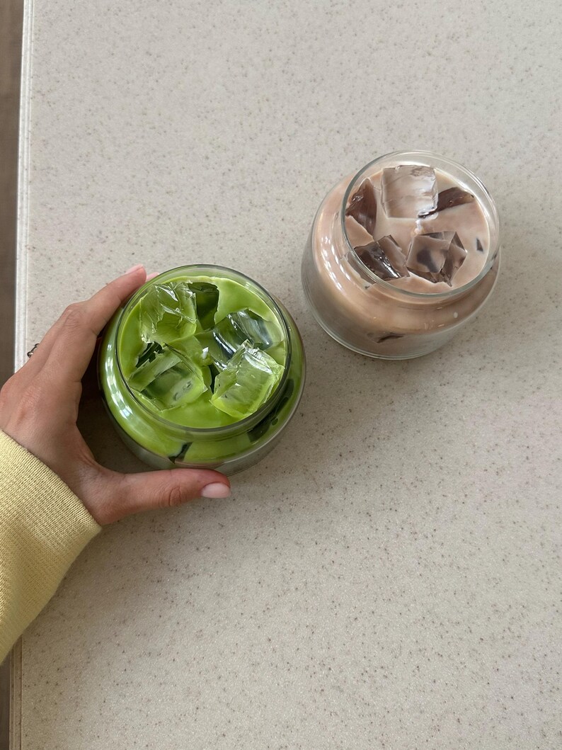 Op de afbeelding: Twee ijskoude drankjes in doorzichtige glazen potten. E&eacute;n is een felgroene matcha latte met ijsblokjes, de andere een lichtbruine koffiedrank met ijs. De drankjes staan op een gespikkeld, lichtgekleurd oppervlak.