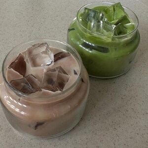 Op de afbeelding: Twee glazen potten gevuld met ijskoude dranken. E&eacute;n pot bevat een lichtbruine drank met ijsblokjes, en de andere een groene drank met ijsblokjes. De potten staan op een gespikkeld, lichtgekleurd oppervlak.
