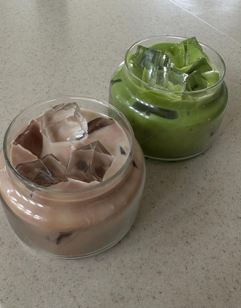 Op de afbeelding: Twee glazen potten gevuld met ijskoude dranken. E&eacute;n pot bevat een lichtbruine drank met ijsblokjes, en de andere een groene drank met ijsblokjes. De potten staan op een gespikkeld, lichtgekleurd oppervlak.