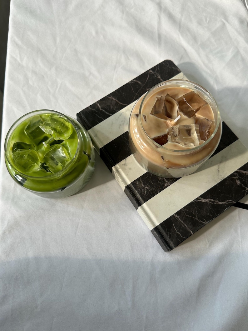 Op de afbeelding: Twee heldere glazen bekers, een gevuld met groene matcha en ijsblokjes, de andere met ijskoffie en ijsblokjes. Ze staan op een zwart-wit gestreept notitieboek, tegen een witte achtergrond.