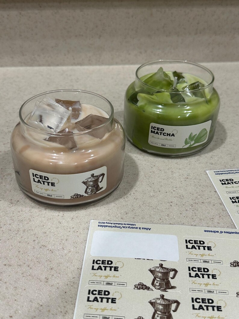 Op de afbeelding: Twee glazen potkaarsen, &eacute;&eacute;n met een lichtbruine tint met het label "ICED LATTE" en de andere groen met het label "ICED MATCHA". Beide kaarsen hebben ijsblokjes. Stickers met het "ICED LATTE" logo zijn ook zichtbaar.