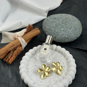 May include: A white ceramic tray with a textured surface and two gold flower-shaped earrings. The tray is sitting on a black surface with a bottle of perfume and cinnamon sticks.