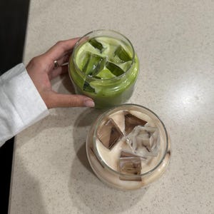 Op de afbeelding: Twee ijskoude drankjes in doorzichtige glazen bekers. E&eacute;n bevat een groene vloeistof met groene ijsblokjes, de andere een lichtbruine vloeistof met bruine ijsblokjes. De drankjes staan op een lichtgekleurd aanrecht.