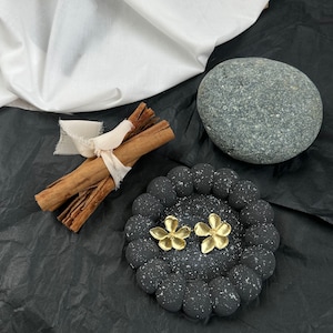 May include: A pair of gold-colored flower earrings displayed on a black, speckled, circular tray. The earrings are the focal point, with a bundle of cinnamon sticks tied with a cream-colored ribbon and a gray stone in the background.