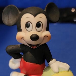 May include: A ceramic figurine of Mickey Mouse, with a black torso, red shorts, and white gloves and shoes. The figure is holding a yellow and green beach ball. The base is yellow.