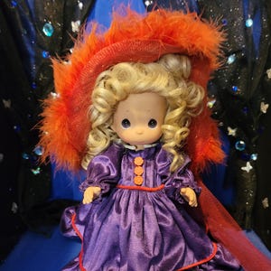 May include: A vintage doll with blonde curly hair, wearing a purple satin dress with orange buttons and trim. The doll is adorned with a large orange feathered hat and a red sheer scarf.
