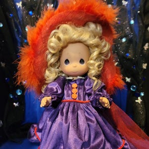 May include: A Precious Moments figurine of a girl with blonde curly hair, wearing a purple satin dress with orange buttons and trim. She is wearing a red hat with orange feathers. The doll has a sweet expression.