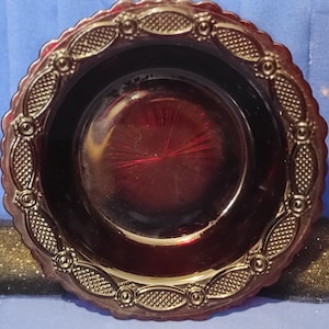 May include: A ruby red glass dish with a decorative rim. The rim features an embossed pattern of oval shapes and small circles. The dish has a scalloped edge and a smooth, reflective surface.