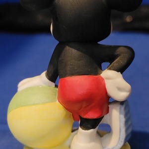 May include: A ceramic Mickey Mouse figurine viewed from the back, wearing red shorts, white gloves, and shoes. He leans on a beach ball with yellow and green sections. The figurine is on a yellow base.