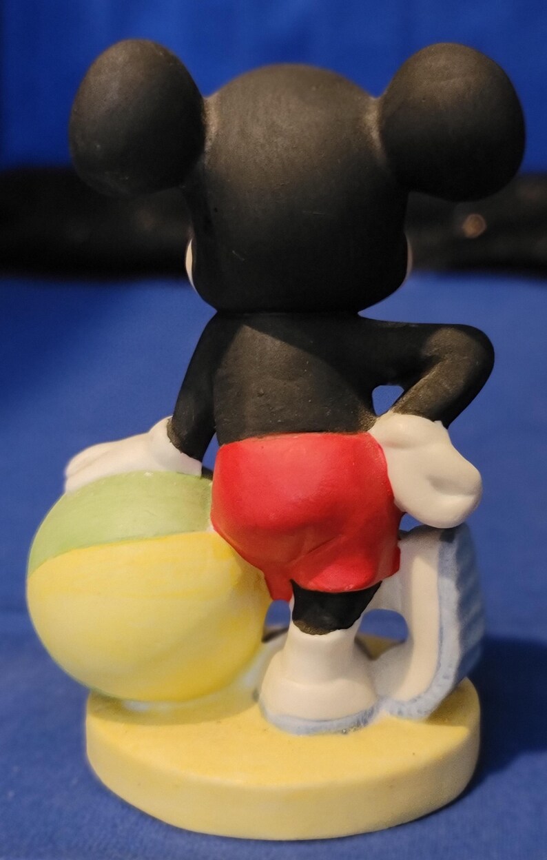 May include: A ceramic Mickey Mouse figurine viewed from the back, wearing red shorts, white gloves, and shoes. He leans on a beach ball with yellow and green sections. The figurine is on a yellow base.
