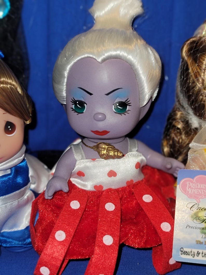 May include: A Precious Moments doll of Ursula from The Little Mermaid. The doll has a purple face, white hair, and a red and white polka dot dress. A gold seashell pendant hangs around her neck. The doll is part of a collection.
