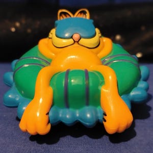 May include: A colorful Garfield figurine in a reclining pose. The figure is primarily orange with a blue hat and green cushion. The cushion has purple stripes. The figurine has yellow whiskers and a pink nose. The overall design is cartoonish and playful.