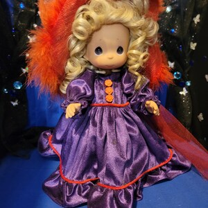 May include: A vintage doll with blonde curly hair, wearing a purple satin dress with orange accents and a red-trimmed hem. The doll is adorned with a large red hat with orange feathers. The doll has a sweet expression and is a collectible item.