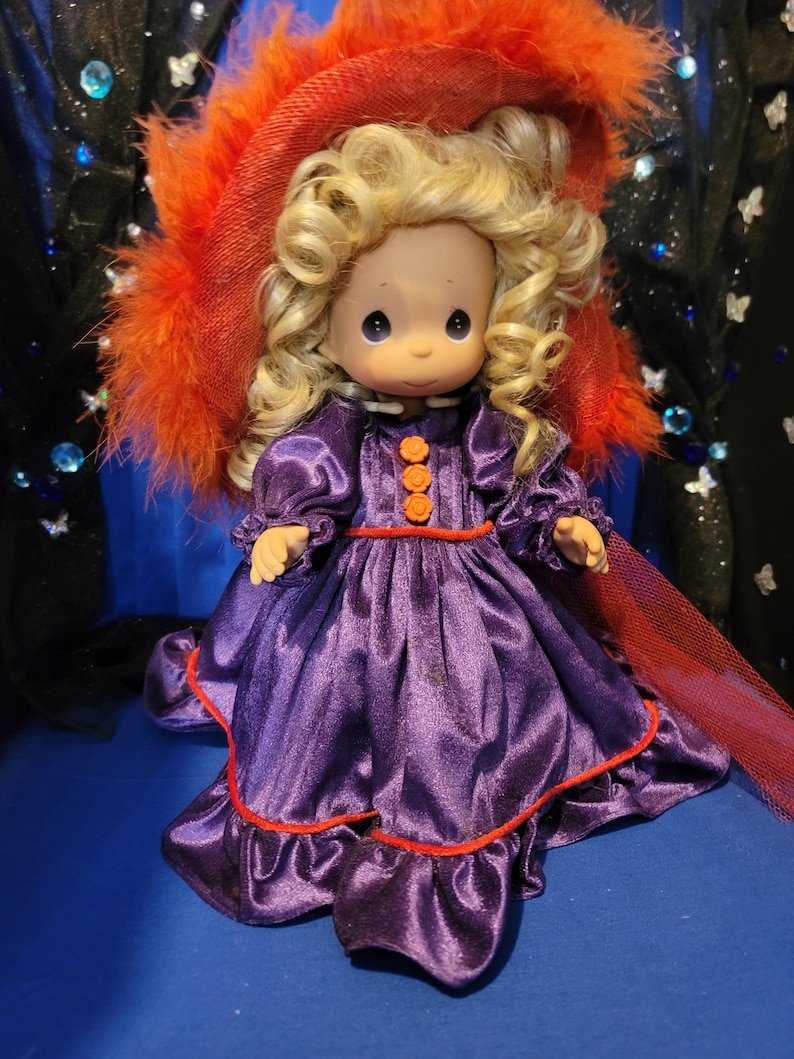 May include: A vintage doll with blonde curly hair, wearing a purple satin dress with orange accents and a red-trimmed hem. The doll is adorned with a large red hat with orange feathers. The doll has a sweet expression and is a collectible item.