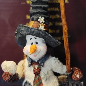 K&ouml;nnte beinhalten: Eine skurrile Schneemannfigur mit schwarzem Hut, Jeansjacke und gestreifter Krawatte. Der Schneemann h&auml;lt einen Besen und einen Schneeball, mit einer Karottennase und einem l&auml;chelnden Gesicht. Sammlerst&uuml;ck.