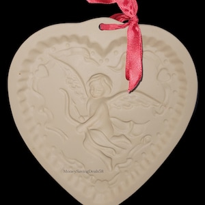 May include: Off-white heart-shaped ornament with an embossed design of Cupid with a bow and arrow. The ornament has a scalloped edge and a red ribbon for hanging. The text "MoneySavingDeals58" is visible on the bottom.
