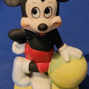 May include: A ceramic figurine of Mickey Mouse holding a yellow and green ball. Mickey is wearing red shorts, a black shirt, white gloves and shoes. The figurine stands on a yellow base. The background is blue.