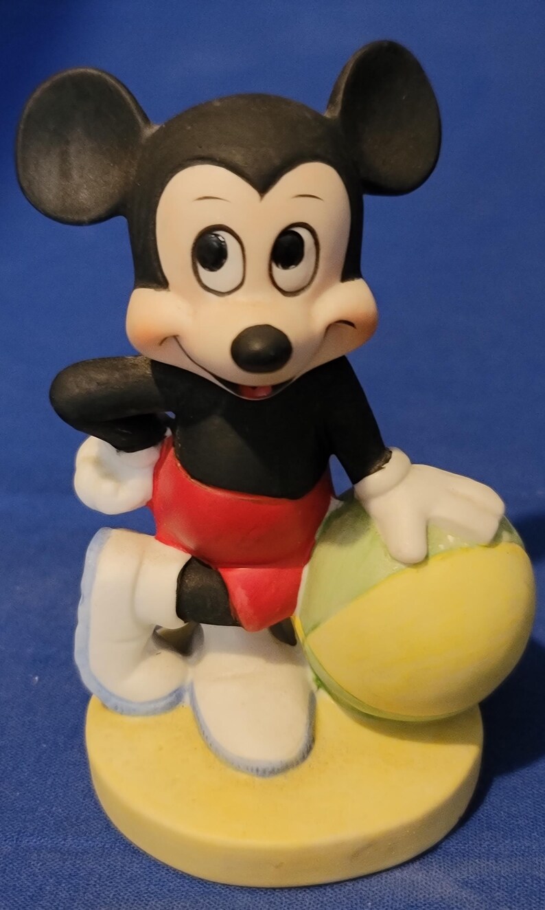 May include: A ceramic figurine of Mickey Mouse holding a yellow and green ball. Mickey is wearing red shorts, a black shirt, white gloves and shoes. The figurine stands on a yellow base. The background is blue.