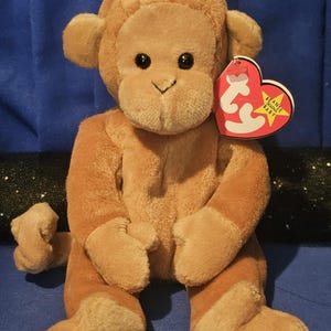 May include: A plush brown monkey toy with a heart-shaped tag that reads "Beanie Baby". The monkey has black button eyes, a tan face, ears, hands, and feet. The toy is sitting on a blue surface.