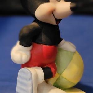 May include: A ceramic figurine of Mickey Mouse holding a beach ball. Mickey is depicted in a black top, red shorts, white gloves, and blue and white shoes. The figurine is on a yellow base. A classic Disney character.