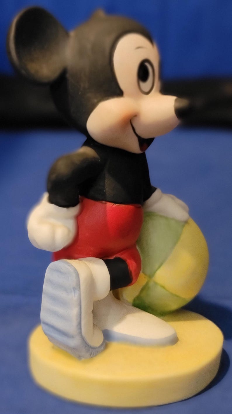 May include: A ceramic figurine of Mickey Mouse holding a beach ball. Mickey is depicted in a black top, red shorts, white gloves, and blue and white shoes. The figurine is on a yellow base. A classic Disney character.