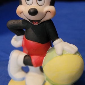 May include: A ceramic Mickey Mouse figurine. Mickey is wearing a black top, red shorts, and white gloves and shoes. He holds a yellow and green ball and stands on a yellow base. The figurine has a smiling expression.