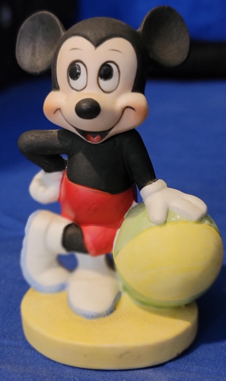 May include: A ceramic Mickey Mouse figurine. Mickey is wearing a black top, red shorts, and white gloves and shoes. He holds a yellow and green ball and stands on a yellow base. The figurine has a smiling expression.