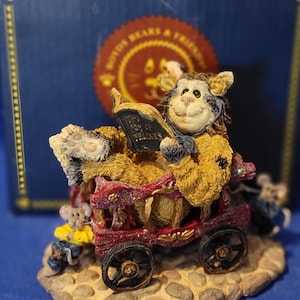 May include: A Boyd's Bears & Friends figurine featuring a smiling bear reading a book while riding in a red wagon. The bear is yellow and brown, with a blue and white face. Two mice are on the wagon and another is in front. The base is stone-like.