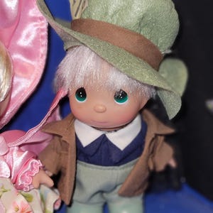 May include: A collectible figurine of a character from Alice in Wonderland, wearing a green hat with a brown band and a tag that reads "10/6". The figurine has a brown jacket, blue shirt, and light green pants. The figurine has white hair and green eyes.