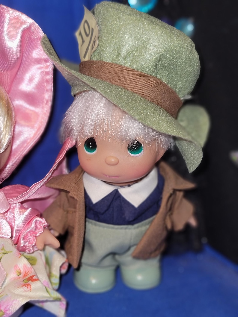 May include: A collectible figurine of a character from Alice in Wonderland, wearing a green hat with a brown band and a tag that reads "10/6". The figurine has a brown jacket, blue shirt, and light green pants. The figurine has white hair and green eyes.