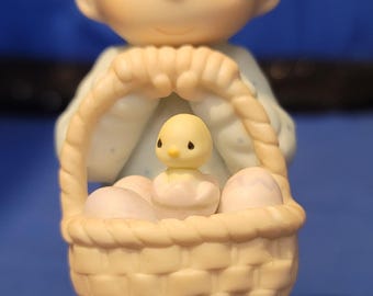 May include: A figurine of a child holding a basket with eggs and a chick. The child has blonde hair and is wearing a light blue outfit. The basket is woven and sits on a patch of grass.