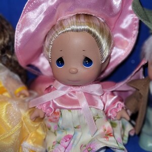 May include: A Precious Moments doll with blonde hair, blue eyes, and a pink bonnet. The doll wears a pink dress with floral print and a matching pink bow. The doll is part of a collection.