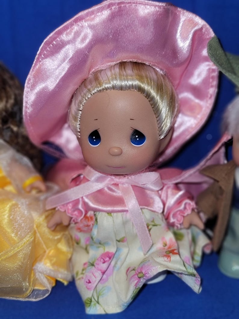May include: A Precious Moments doll with blonde hair, blue eyes, and a pink bonnet. The doll wears a pink dress with floral print and a matching pink bow. The doll is part of a collection.