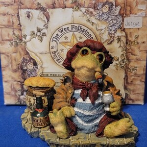 May include: A figurine of a frog dressed as a wine taster, holding a glass and a cigar. The frog wears a brown hat, jacket, and a blue and white striped shirt. The base reads "THE WINE TASTER". A bottle of "OLD TOAD" is also present.