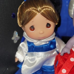 May include: A porcelain doll with brown hair and eyes, wearing a white and blue outfit with a matching blue bow. The doll has a sweet expression and is part of a collectible figurine series.