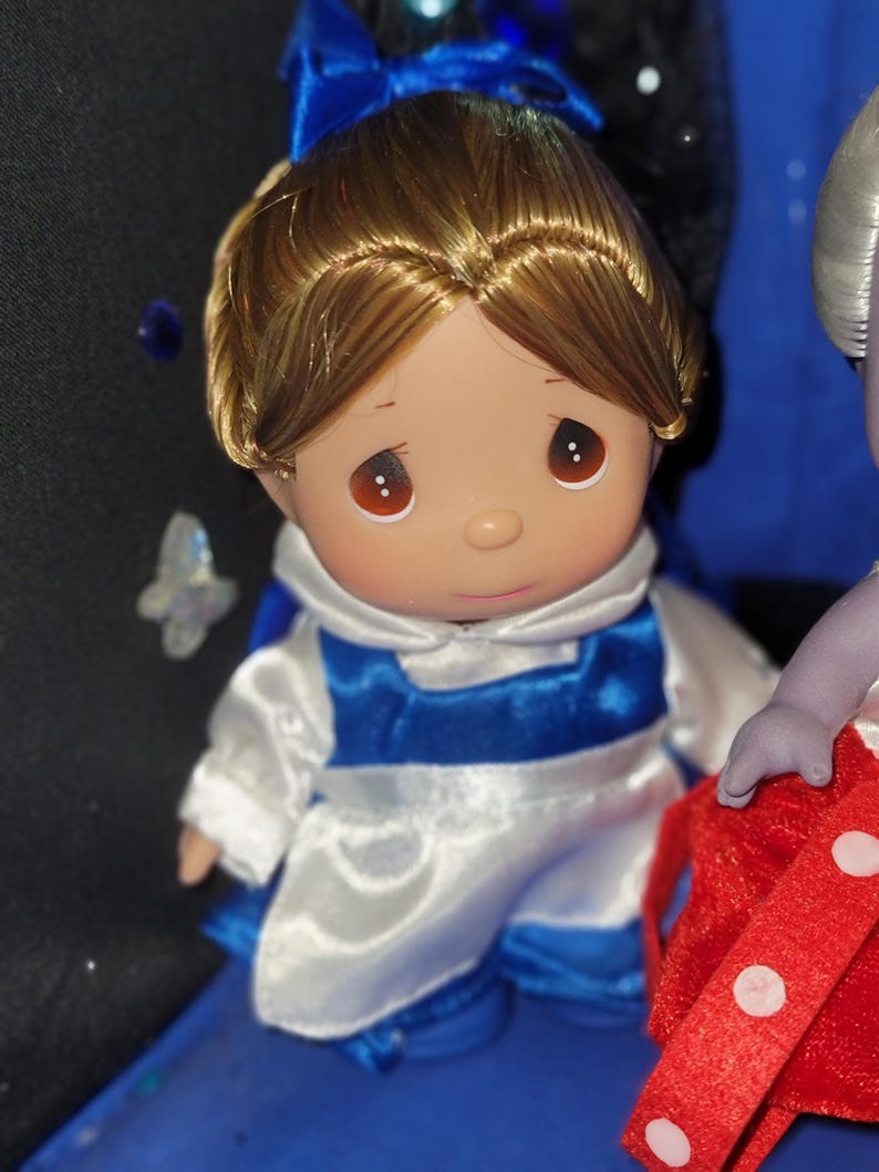 May include: A porcelain doll with brown hair and eyes, wearing a white and blue outfit with a matching blue bow. The doll has a sweet expression and is part of a collectible figurine series.