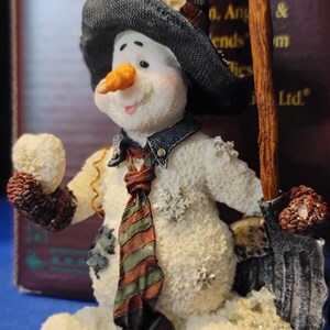 K&ouml;nnte beinhalten: Eine skurrile Schneemannfigur mit schwarzem Hut, Jeansweste und gestreifter Krawatte. Der Schneemann hat eine Karottennase, ein l&auml;chelndes Gesicht und h&auml;lt einen Besen. Die Figur ist mit Schneeflocken verziert und hat ein strukturiertes, schneegleiches Aussehen.