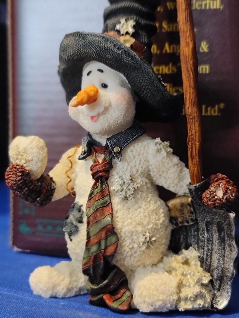 K&ouml;nnte beinhalten: Eine skurrile Schneemannfigur mit schwarzem Hut, Jeansweste und gestreifter Krawatte. Der Schneemann hat eine Karottennase, ein l&auml;chelndes Gesicht und h&auml;lt einen Besen. Die Figur ist mit Schneeflocken verziert und hat ein strukturiertes, schneegleiches Aussehen.