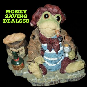 May include: A whimsical frog figurine, "The Wine Taster," is depicted wearing a brown jacket, red scarf, and a blue and white striped shirt. The frog holds a wine glass and a lit cigar. A barrel, bottle, and bread are also present.