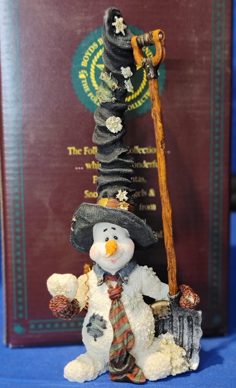 K&ouml;nnte beinhalten: Eine skurrile Schneemannfigur mit einem hohen schwarzen Hut, der mit wei&szlig;en Akzenten verziert ist, und einer passenden Schaufel. Der Schneemann tr&auml;gt eine gemusterte Krawatte und h&auml;lt einen kleinen Gegenstand. Teil der "The Folktales Collection".