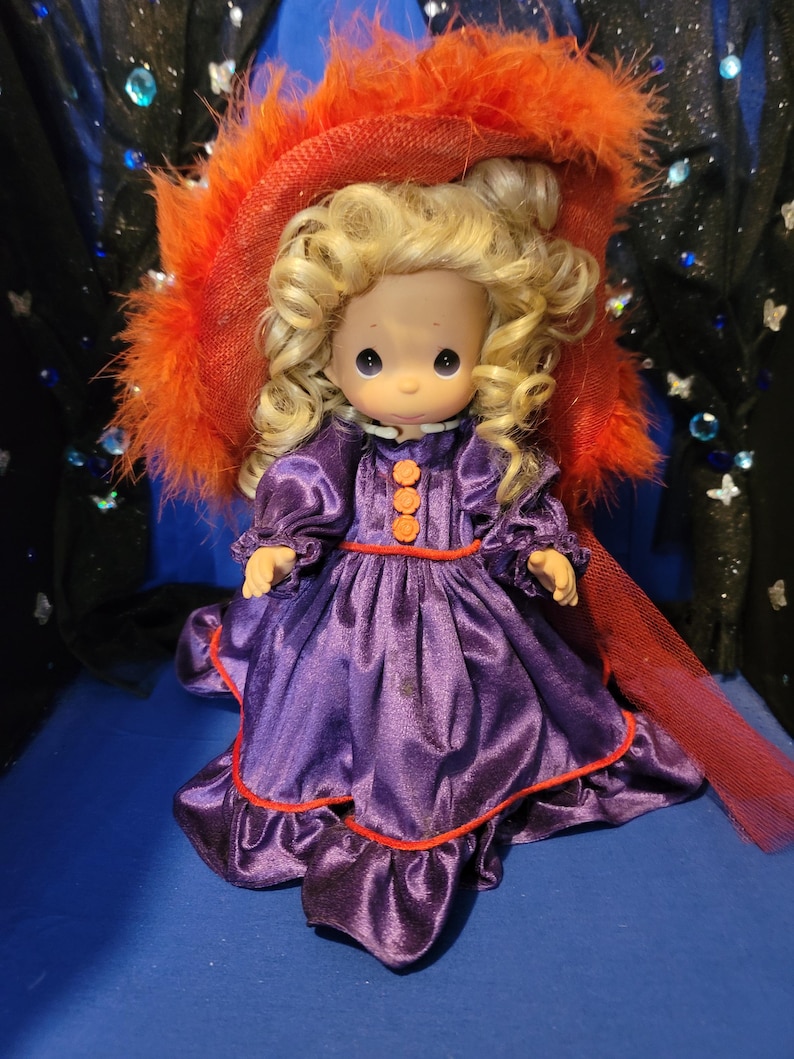 May include: A vintage doll with blonde curly hair, wearing a purple satin dress with orange buttons and trim. The doll is adorned with a large orange feathered hat and a red sheer scarf.