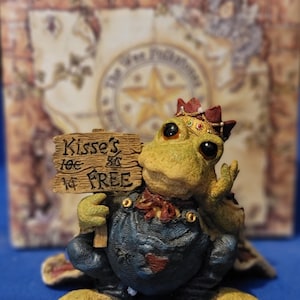 May include: A whimsical figurine of a turtle wearing a crown and overalls, holding a sign that reads "Kisses 10¢, 1¢ FREE". The turtle is green and yellow, with a red and gold crown. The background is a blurred image of a map.