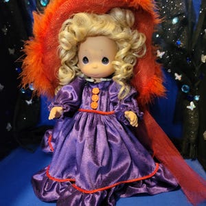 May include: A Precious Moments doll wearing a purple satin dress with orange trim and buttons. The doll has blonde curly hair and is wearing a large orange hat with feather trim. The doll is a collectible item.