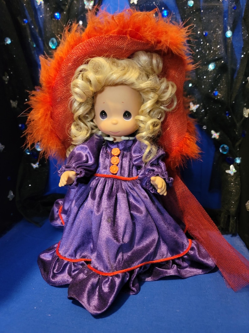 May include: A Precious Moments doll wearing a purple satin dress with orange trim and buttons. The doll has blonde curly hair and is wearing a large orange hat with feather trim. The doll is a collectible item.